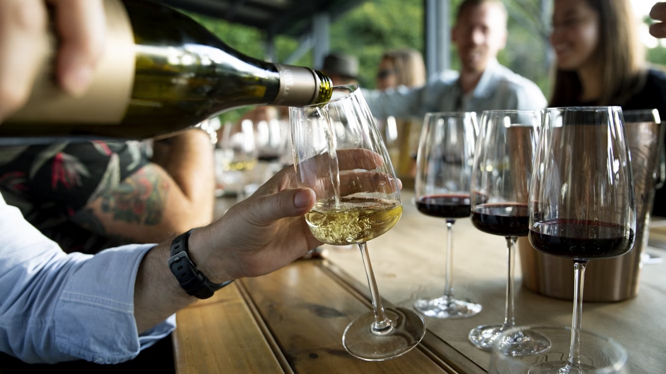 Mãos segurando uma garrafa de vinho branco servindo uma taça em uma mesa com outras taças de vinho tinto e pessoas ao fundo, sugerindo uma degustação de vinhos ou aprendizado sobre vinhos em grupo.