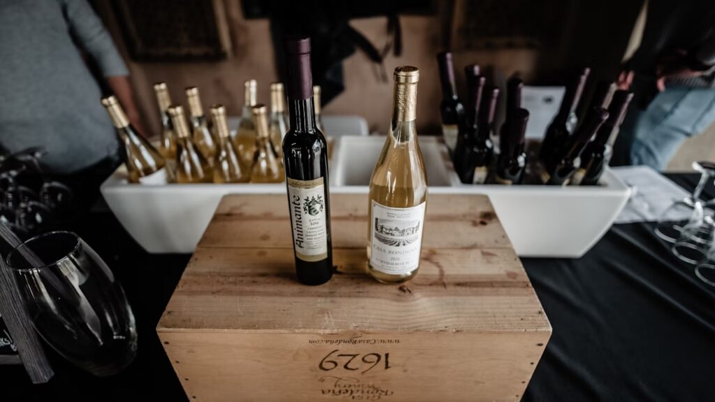 Garrafas de vinho branco e tinto expostas, recebidas de clube do vinho, com destaque para duas garrafas sobre uma caixa de madeira.